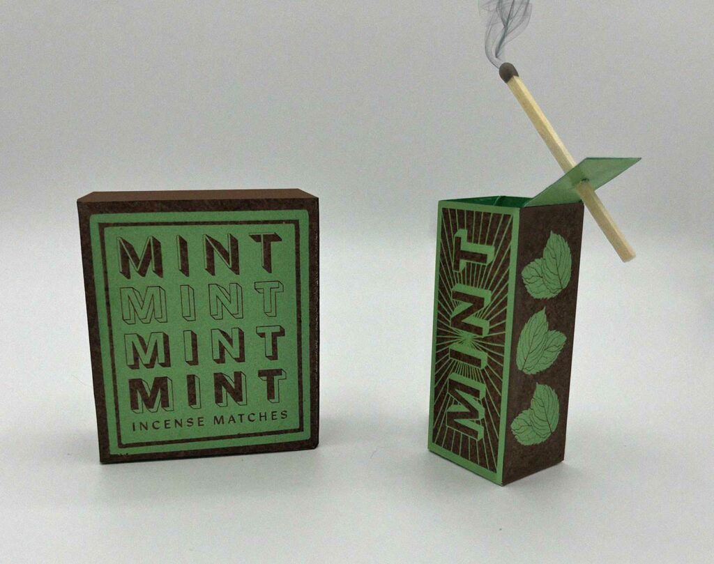 Photo of the mint match boxes and next to it is the match holder with a match in it, lit up.