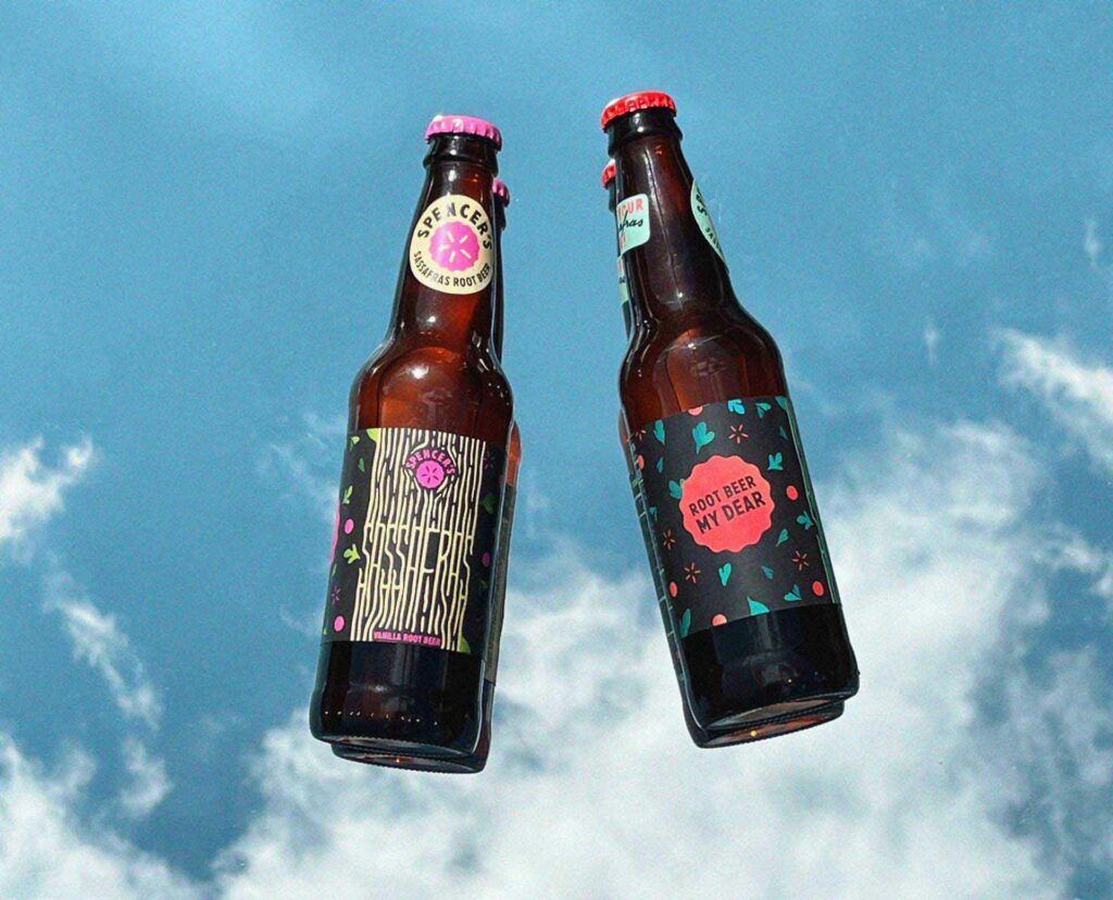 Two root beer bottles, one classic and one vanilla, laying on a mirror background with a partially blue sky with clouds as the background.