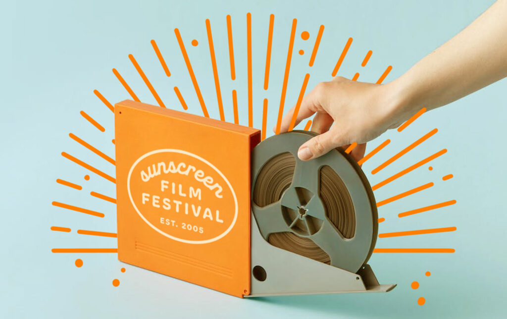 A photo of a hand reaching out to a film reel emerging from a film reel box. On the side of the box is the Sunscreen Film Festival logo. Illustrated yellow sun rays are surrounding the reel.